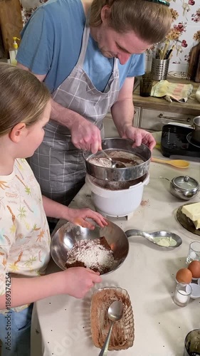 Young dad in apron and his young daughter cooking together in the kitchen indoors. They share family leisure time, enjoying food preparation and bonding through fun interaction in a warm home setting