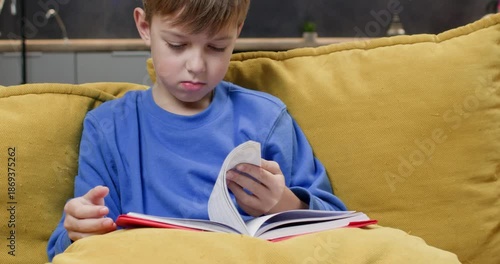 Calm schoolboy turns pages of book sitting on yellow sofa cushions. Preteen child reads interesting story resting in cozy domestic atmosphere. Education