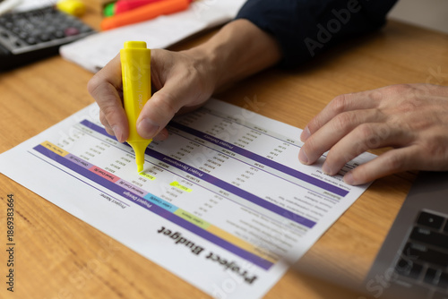 Person highlighting figures on project budget sheet with yellow marker, analyzing financial data and expenses. Concept for budgeting, accounting, financial planning business cost control.