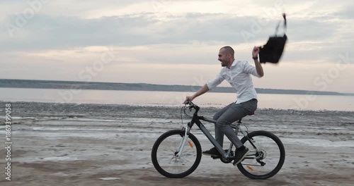 Playful businessman cycles along wet sea coastline swinging briefcase energetically in warm wind. Cheerful man enjoys rhythmic motion under glowing seaside sky