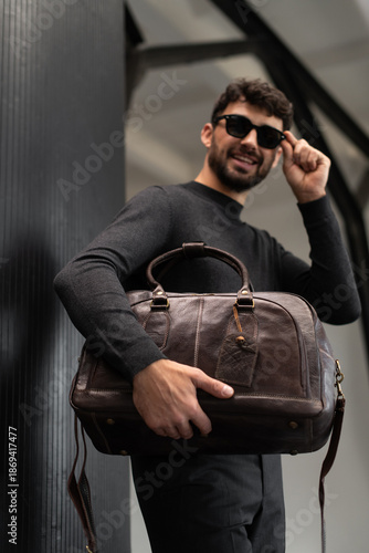 Confident man in sunglasses and charcoal knitwear holding a dark brown leather briefcase while leaning against a textured pillar in a modern minimalist interior.