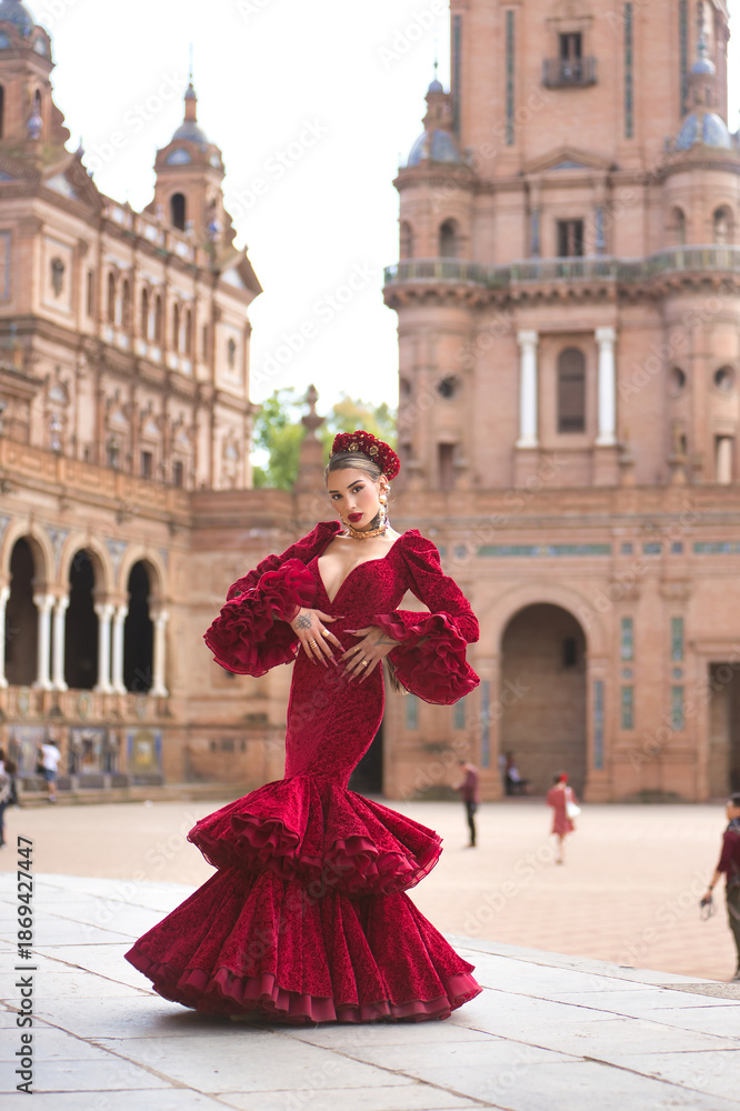 Naklejka premium Young, beautiful, blonde woman in red flamenco dress, flowers in her hair and gold jewelry, performing flamenco poses in the square of Spain in Seville. Dance concept, art. Vertical position.
