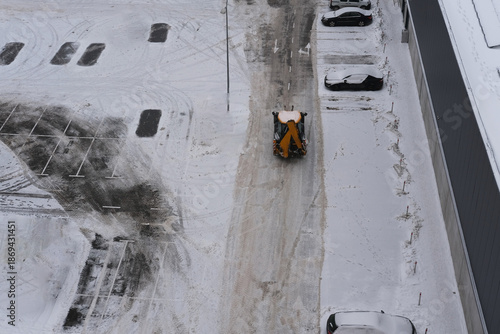 Snow plow clearing a parking lot covered in fresh snow, with tire tracks visible and parked vehicles in the background, showcasing winter maintenance and urban landscape