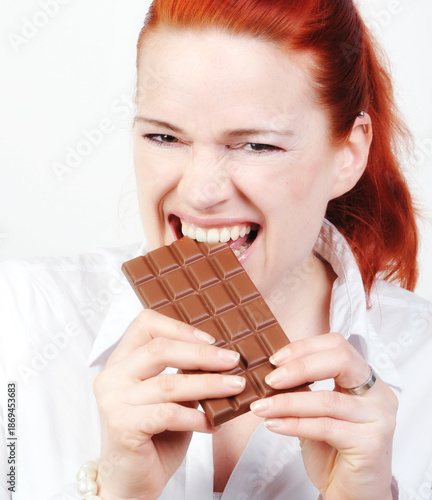 Junge Frau mit roten Haaren beißt genussvoll in eine Tafel Schokolade vor hellem Hintergrund. Das Motiv steht für Genuss, Lust, Heißhunger, Süßigkeiten und emotionale Ernährung.