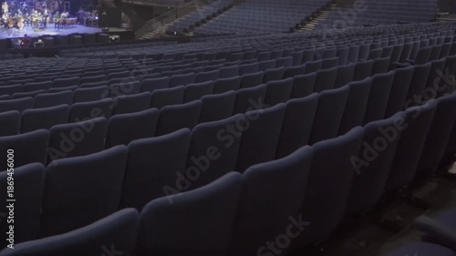 Auditorium filled with rows of empty seats near brightly lit stage with orchestra. Atmosphere of event rehearsal with focus on performers