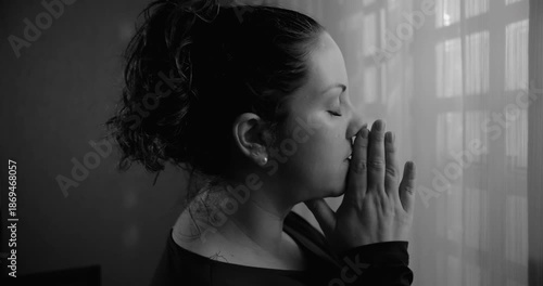 Woman standing by window in monochrome with hands pressed together in prayer, deep contemplation, spiritual focus, quiet surrender, intimate moment of faith