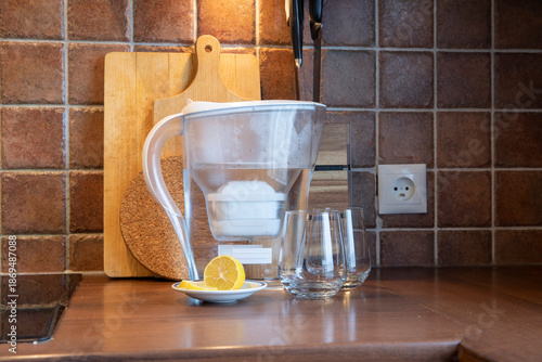 Water filter pitcher with clean drinking water, empty glasses and fresh lemon on a kitchen countertop. Concept of water purification, healthy lifestyle and home hydration, domestic use.