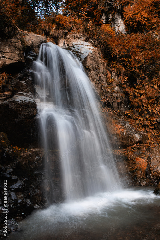 Obraz premium Water cascades down a rocky surface in a forest. Autumn leaves surround the waterfall, creating a vibrant natural scene in the afternoon sun.