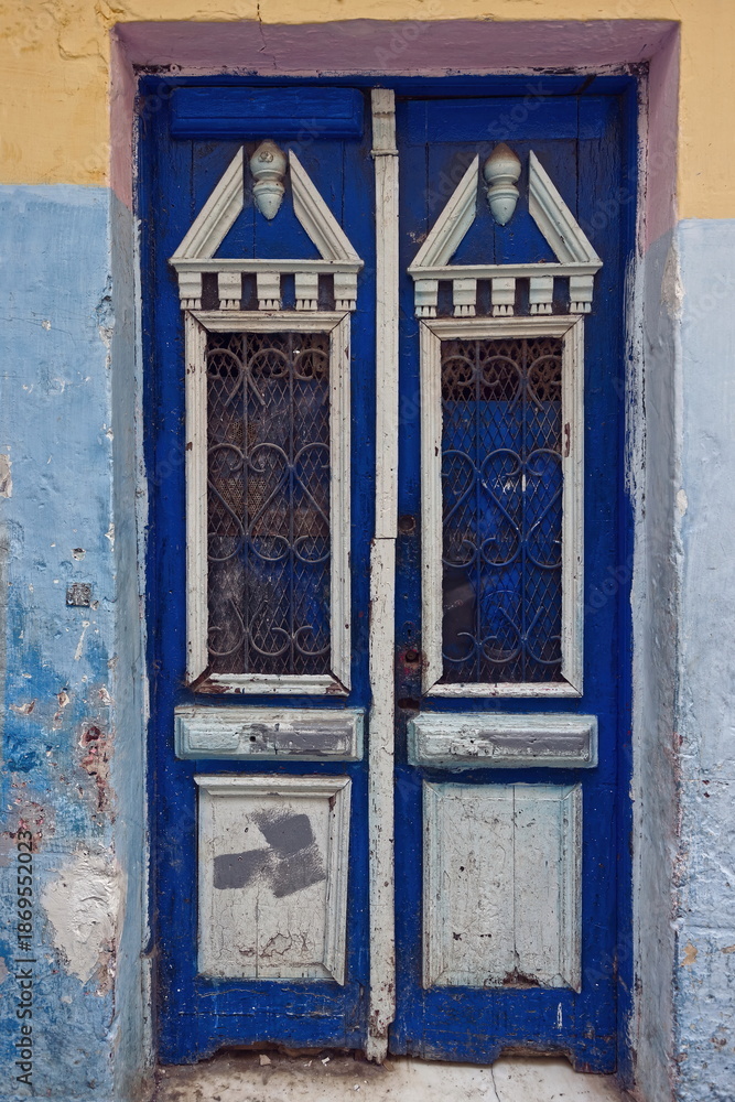 Naklejka premium Wooden door haphazardly painted in indigo blue and white with neo-Classical ornate, on a mustard, pink and light blue faded wall. Tangier-Morocco-009