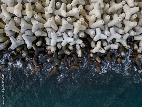Top-down aerial view of massive concrete wave breakers meeting the sea, showing contrast between rigid human-made infrastructure and powerful natural water, forming an abstract, graphic coastal