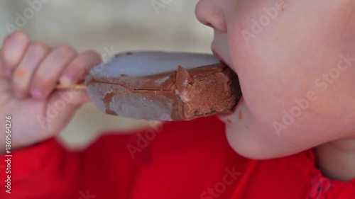 Tight closeup of a small child in a red jacket biting a chocolate ice cream bar on a wooden stick. Appetite and sweet treat concept, food and childhood, copy space