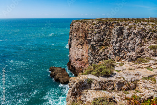 A stunning close-up view of the imposing cliffs at Cabo de São Vicente in Sagres, Portugal, where jagged rock formations plunge dramatically into the deep blue Atlantic Ocean.