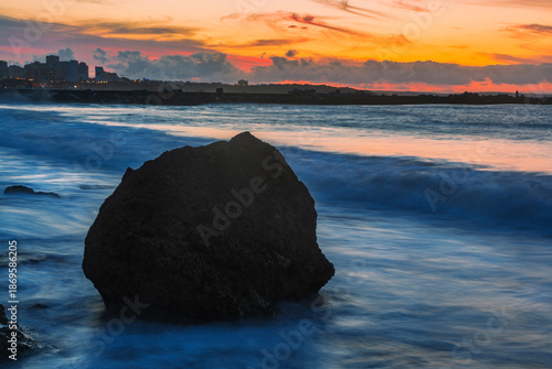A breathtaking sunset view from Praia Pintadinho in the Algarve, Portugal, where a solitary, dark rock stands silhouetted against the vibrant orange and pink sky.