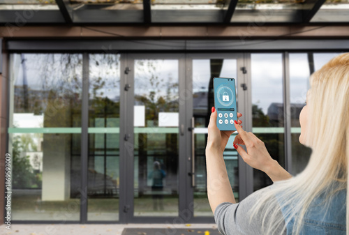 Female hand holding black mobile smart phone with smart home application on the screen. Blurred house on the background. For access to all of the controls of your house and caring of home security