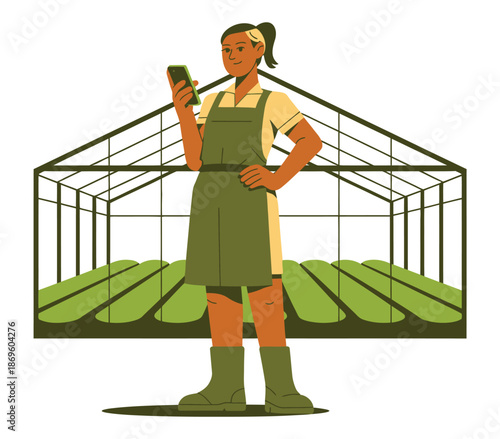 A woman in an apron uses a smartphone in front of a greenhouse with rows of plants.