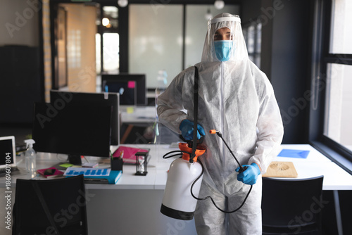 Male technician wearing coveralls and face shield, spraying disinfectant with sprayer in office