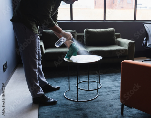 Man wearing gloves cleaning side table in office lounge with spray bottle and cloth, copy space