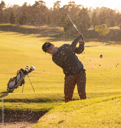 Male golfer swinging club, hitting golf ball from sand bunker on golf course near golf bag