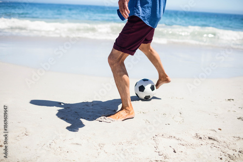 Adult male kicking black-and-white soccer ball on sandy ocean shore with gentle waves lapping