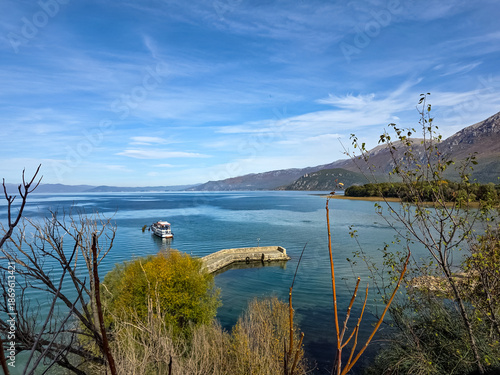 Fotografía Scenic view of Lake Ohrid featuring a small boat near a pier, calm clear water, surrounding mountains, and blue sky, showcasing peaceful nature and travel destination in Ohrid region