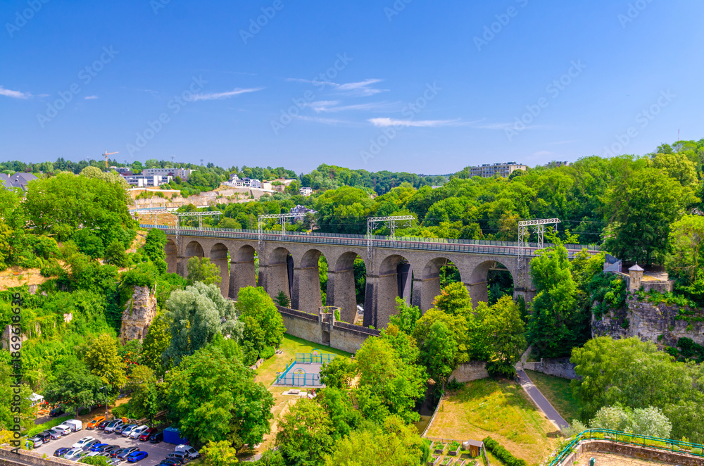 Fototapeta premium Pulvermuhl Viaduct Biisser Breck railway stone arch viaduct bridge across Alzette valley and Maierchen stone defensive wall in Luxembourg City centre, park with green trees and blue sky background
