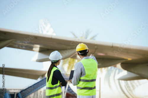 Fotografi Two engineers in safety helmets and reflective vests inspect an aircraft landing gear tire while reviewing maintenance data on digital tablets at an airport maintenance area