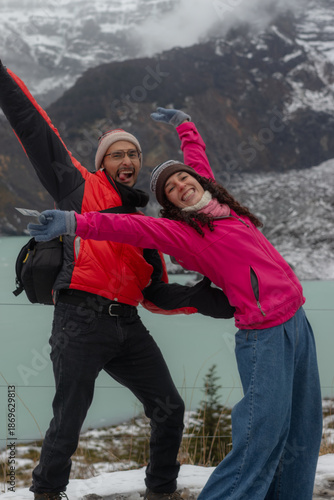 Portrait of a hispanic man and woman travelers posing playfully with arms raised and sticking tongue out near a glacier lake. Concept of friendship, fun and winter tourism.