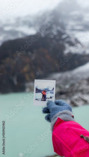 Point of view shot of a gloved hand holding a printed instant photograph of a couple hugging, against a blurred background of a glacier and mountains. Concept of travel memories and souvenirs.