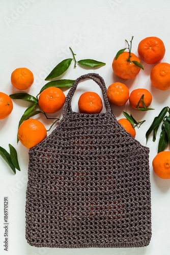Vibrant orange tangerines with fresh green leaves in a reusable crochet bag on a white background, highlighting natural freshness and healthy eating. Copy space.