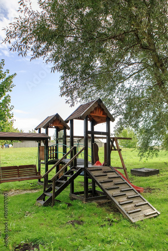 Fotografie Children's outdoor playground made of brown wood with a slide and climbing ramp, located in a peaceful green park environment
