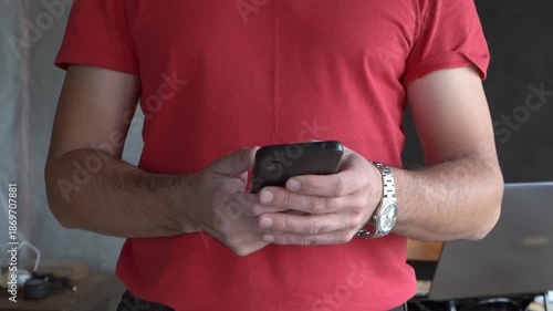 A man holding a smartphone. Holding a phone in his hand. Red T-shirt. Close-up. Writing a message.