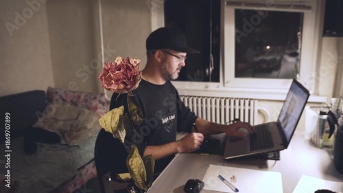A man in black clothes sits at a table. He wears a baseball cap and glasses. He works remotely. Freelancer. Dim lighting. Vintage style. Room. Dried rose. Close-up.