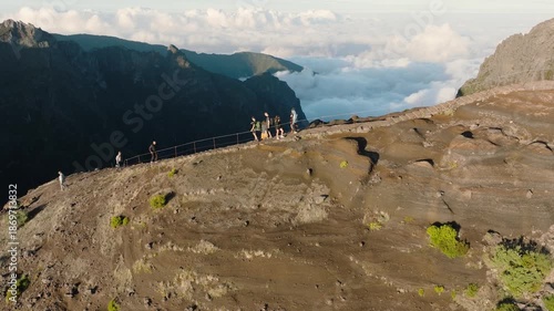 Cinematic side-view aerial drone shot capturing hikers walking along the exposed mountain ridge on famous trail from Pico do Arieiro to Pico Ruivo. The camera follows the ridge from the sunlit side 