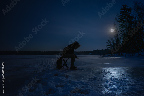 Fotografie Silhouette of a person ice fishing on a frozen lake at night under moonlight, Finland