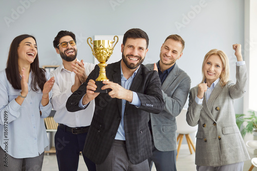 Cuadro en lienzo Portrait of a happy smiling young man winner holding the trophy award for success in business standing in office with a team of people colleagues applauding for company best employee reward