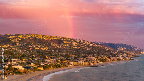 Aerial view of a rainbow arching over the coastal homes and sandy beach, the sky painted with pastel hues, as the ocean meets the shore, Laguna Beach, California, United States.