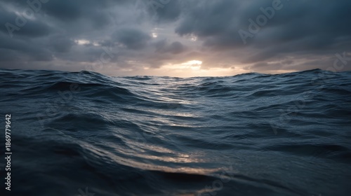 A dramatic moody sunset unfolds over the vast choppy ocean surface with golden light breaking through heavy overcast clouds reflecting on the water