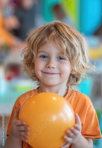 A young boy with blonde, curly hair smiles directly at the camera, holding a bright orange balloon. He wears a simple orange t-shirt. 