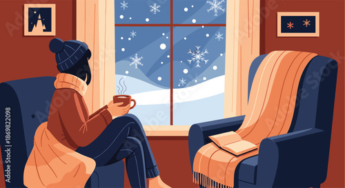 Cozy scene of a woman looking out a window at a snowy night while sitting in a dark blue armchair with a warm drink.
