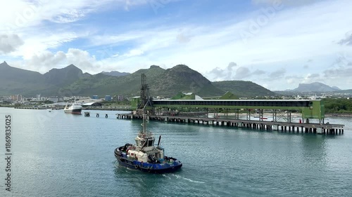 A tugboat works in a busy waterway close to the city harbor. It navigates the water as ships await assistance. Workers are seen on board, managing operations.