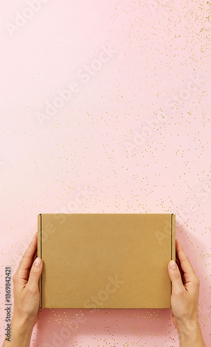 Female hands holding a plain cardboard box on pink background with copy space. Concept of delivery, packaging, online shopping, e-commerce, gift, subscription box, business presentation. 