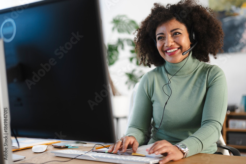A smiling woman wearing a headset works at her computer, typing on the keyboard and looking at the screen.