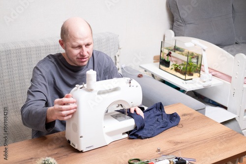 A bald man of 40 years old in a gray T-shirt sits at a table and sews on a sewing machine at home