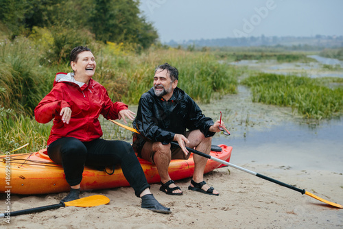 Billede på lærred Happy senior couple relaxing by kayak in rain