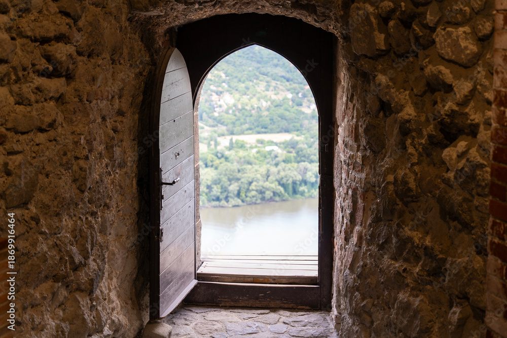 Obraz premium Old wooden door opening into nothingness in a historic castle on a mountaintop, with a river and forest landscape in the distance
