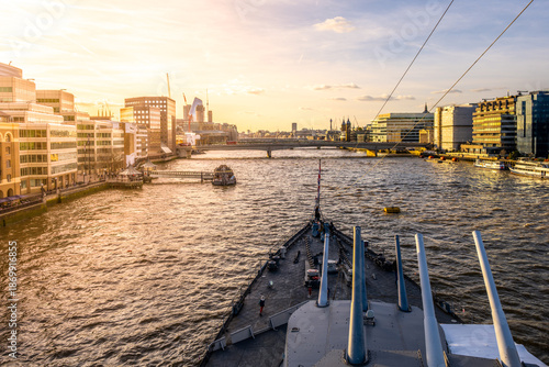 Turrets stand tall on HMS Belfast as the sun sets behind the London skyline. The River Thames flows nearby, with city life bustling along the waterfront.