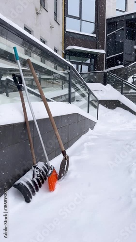 Snow Removal Tools Including Shovels and a Broom Leaning Against Building Railing During Heavy Snowfall, Vertical footage of Winter Maintenance Equipment Prepared for Work.Snow Cleaning Equipment Set