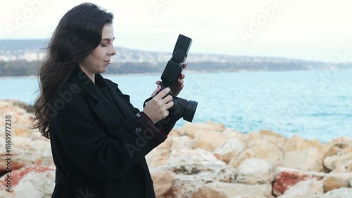 Smiling female photographer holding a camera on a sunny beach, enjoying golden hour light and capturing outdoor moments on the coastline.