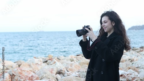 Smiling female photographer holding a camera on a sunny beach, enjoying golden hour light and capturing outdoor moments on the coastline.