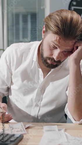 Man sits at a desk reviewing bills with a calculator in a bright office setting.